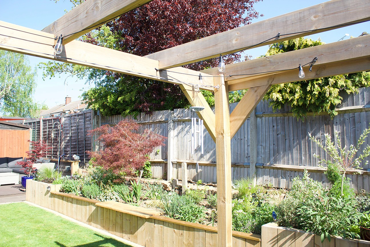 A wooden pergola in a bright garden with various plants and green foliage under a clear blue sky.