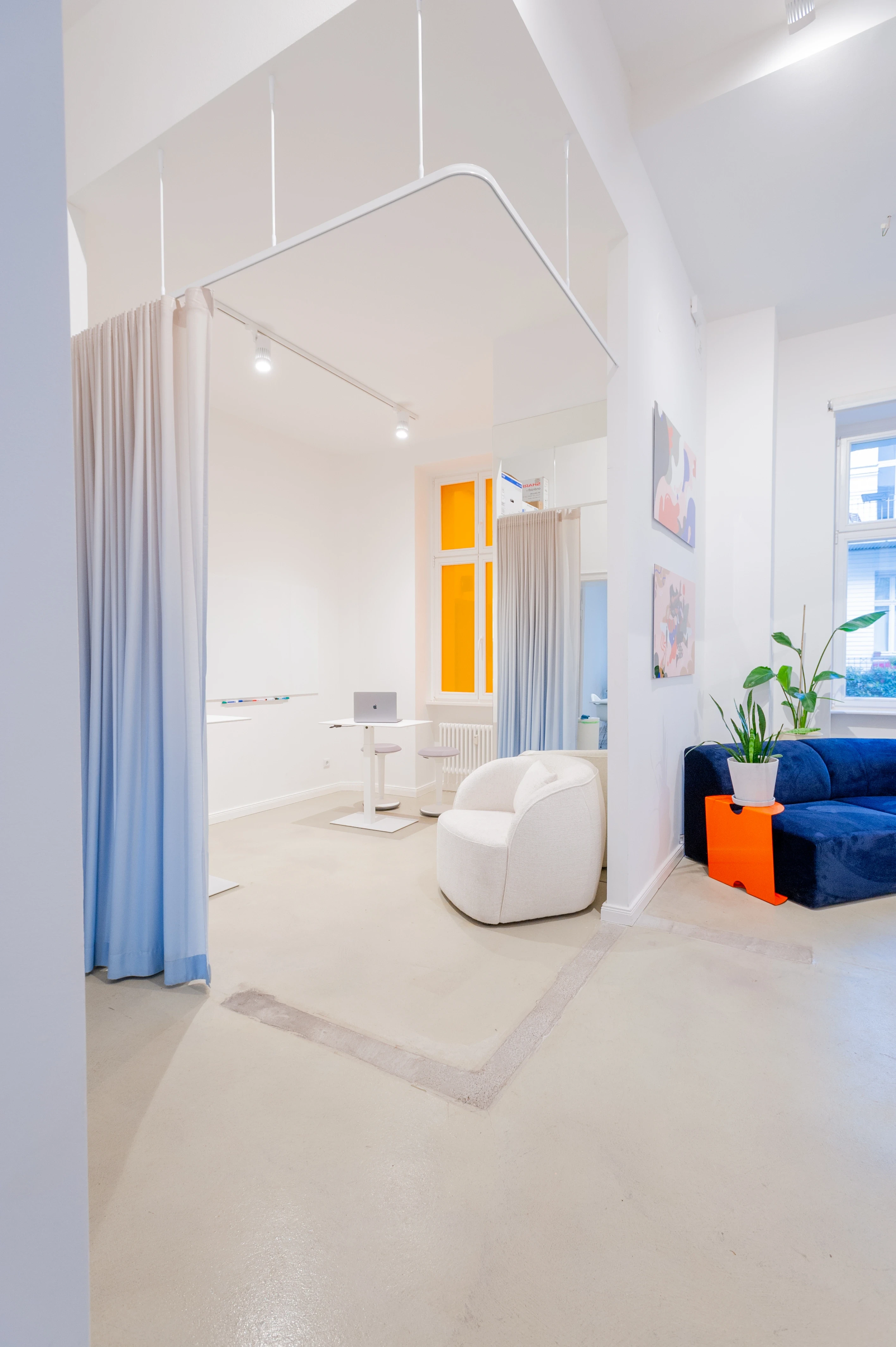 White studio space at BATTLE ROYAL with a curtained focus zone in pale blue, a white rounded armchair, a navy velvet sofa with orange side table, an orange window frame and a potted plant.