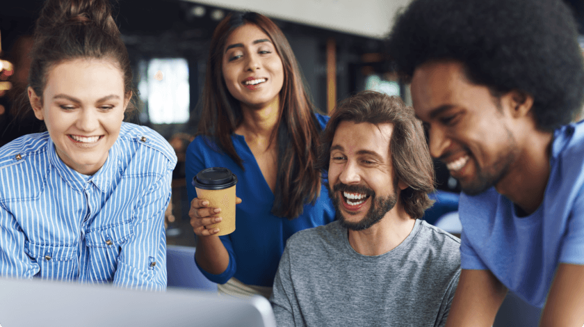 A diverse group of four smiling people gather around a laptop, with one person holding a coffee cup, suggesting a collaborative work environment in a casual office setting.