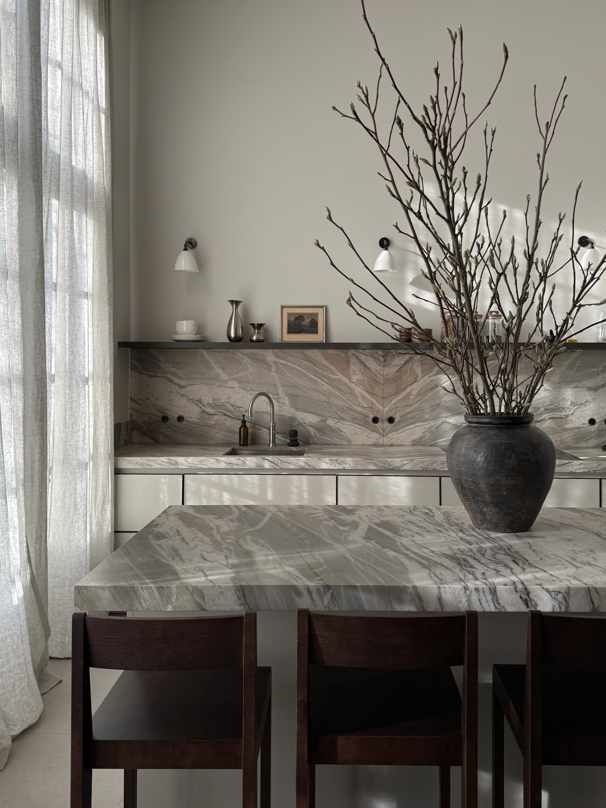 Modern kitchen with book-matched quartzite island and backsplash, weathered ceramic vase with Magnolia branches, and dark walnut bar stools