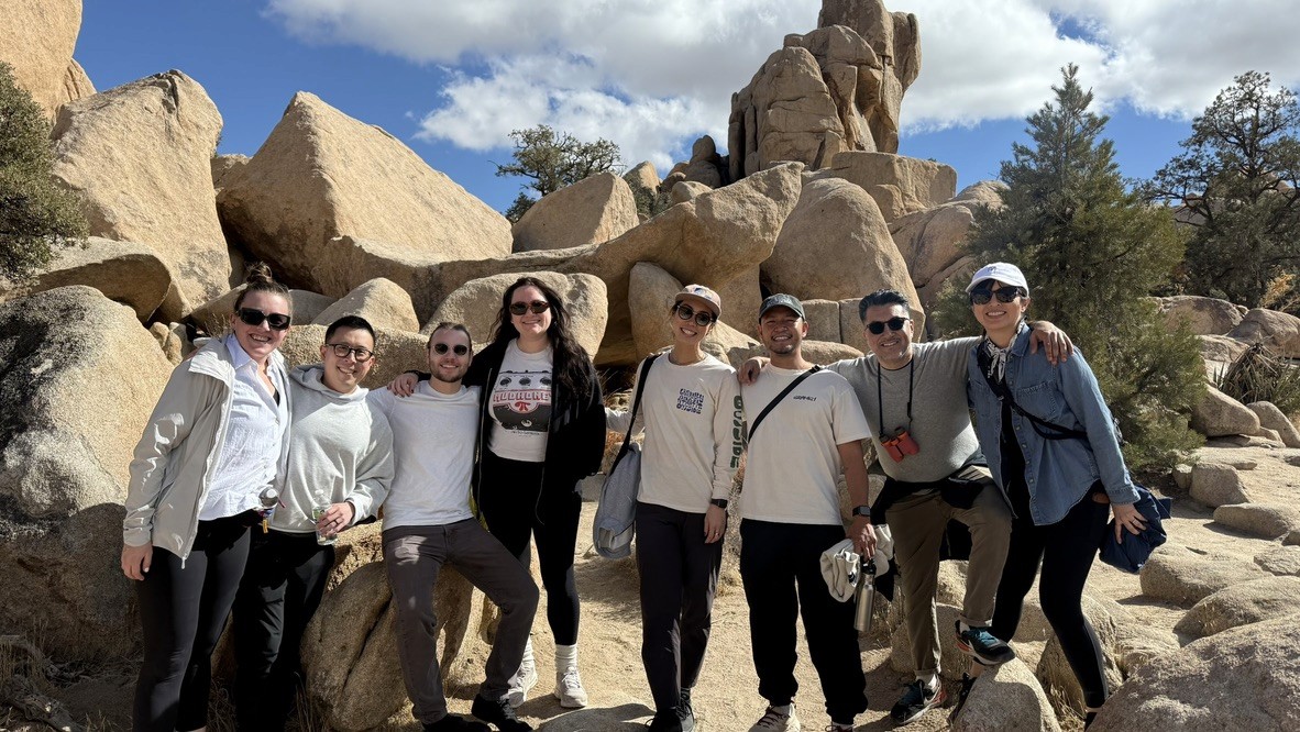Group of people standing together and smiling in front of large desert rock formations on a sunny day.