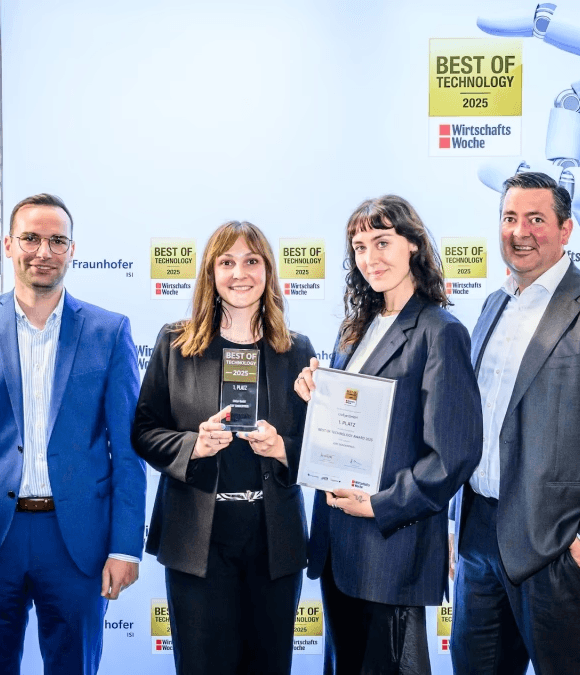 Unser Team steht vor einer Sponsorenwand, die mit dem Logo 'BEST OF TECHNOLOGY 2025' und 'WirtschaftsWoche' bedruckt ist. Die beiden Frauen in der Mitte halten jeweils eine Auszeichnung in die Kamera: eine hält einen dunklen Pokal mit der Aufschrift '1. PLATZ', die andere hält eine gerahmte Urkunde, ebenfalls mit der Auszeichnung '1. PLATZ'. Alle vier Personen lächeln und tragen formelle Kleidung (Anzüge und Business-Kleidung).