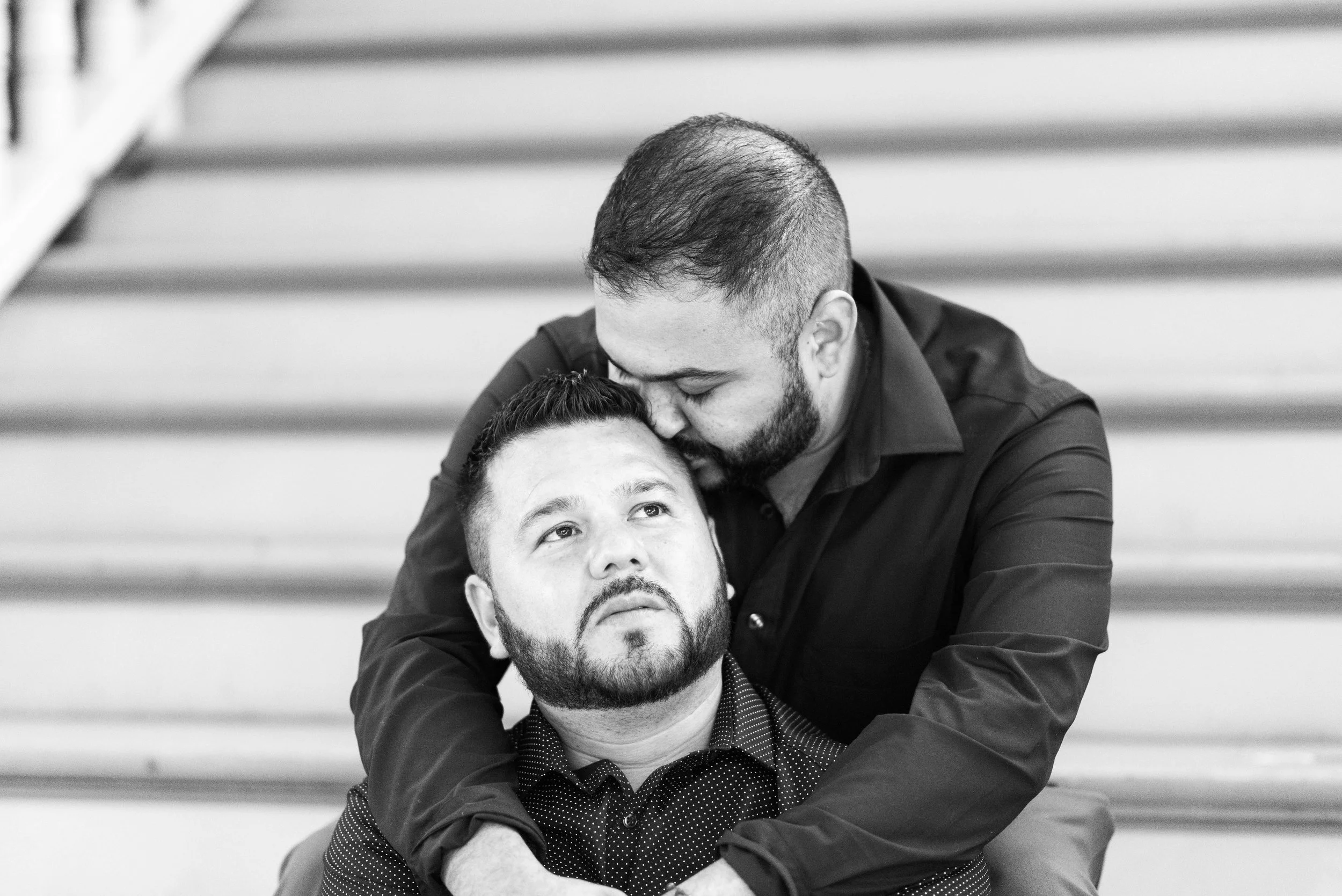 Duas pessoas compartilham um momento íntimo e afetuoso em uma escadaria, com uma abraçando a outra por trás, ambas vestindo camisas escuras, transmitindo uma sensação de acolhimento e conexão.