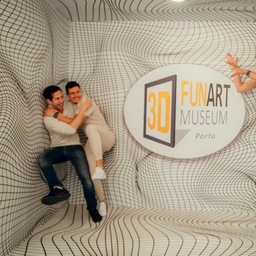 Two people pose against a wall with a 3D illusion background at the 3D Fun Art Museum in Porto. Only arms of a third person are visible.