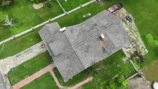 Aerial view of a single-story house with a gray roof, surrounded by green grass and nearby structures.