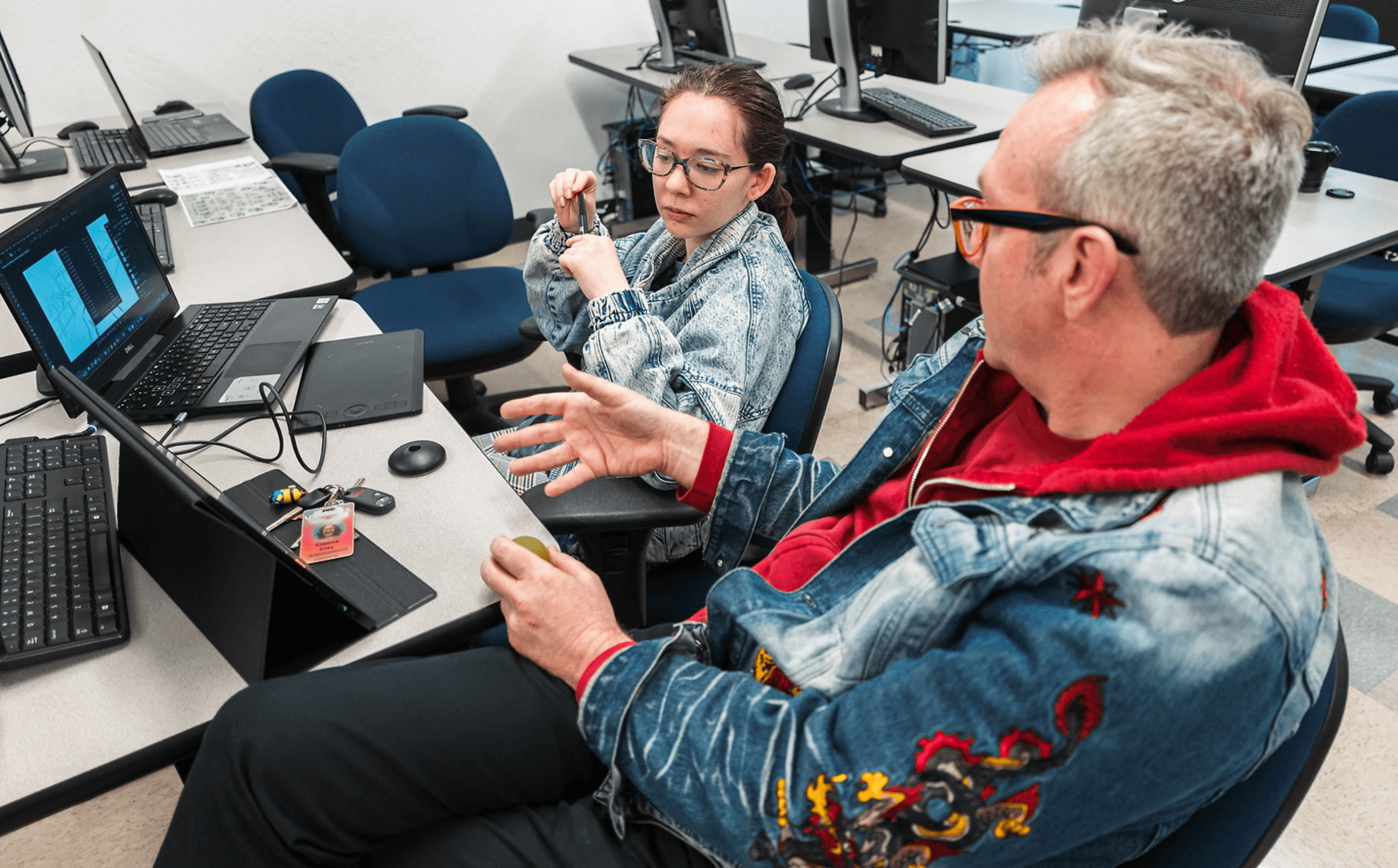 Two people in a computer lab engage in discussion, each wearing denim jackets with colorful designs, surrounded by laptops and electronic equipment.