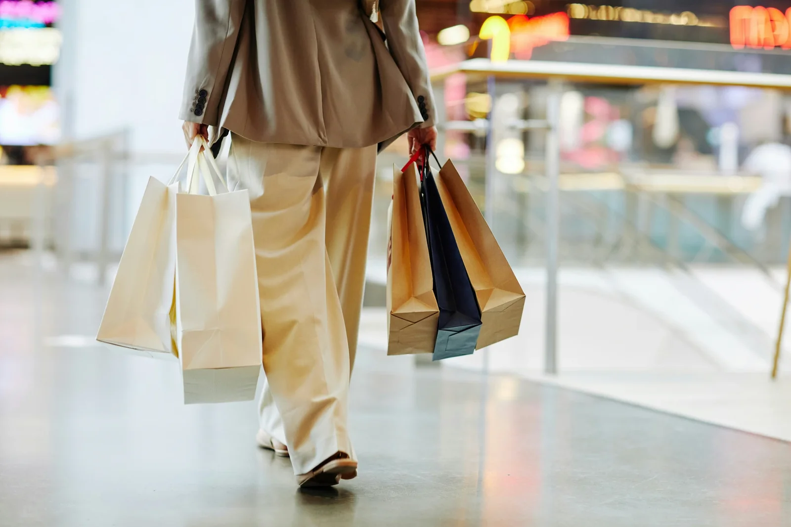 Shopper carrying multiple paper shopping bags walks through a blurred mall interior with storefronts.