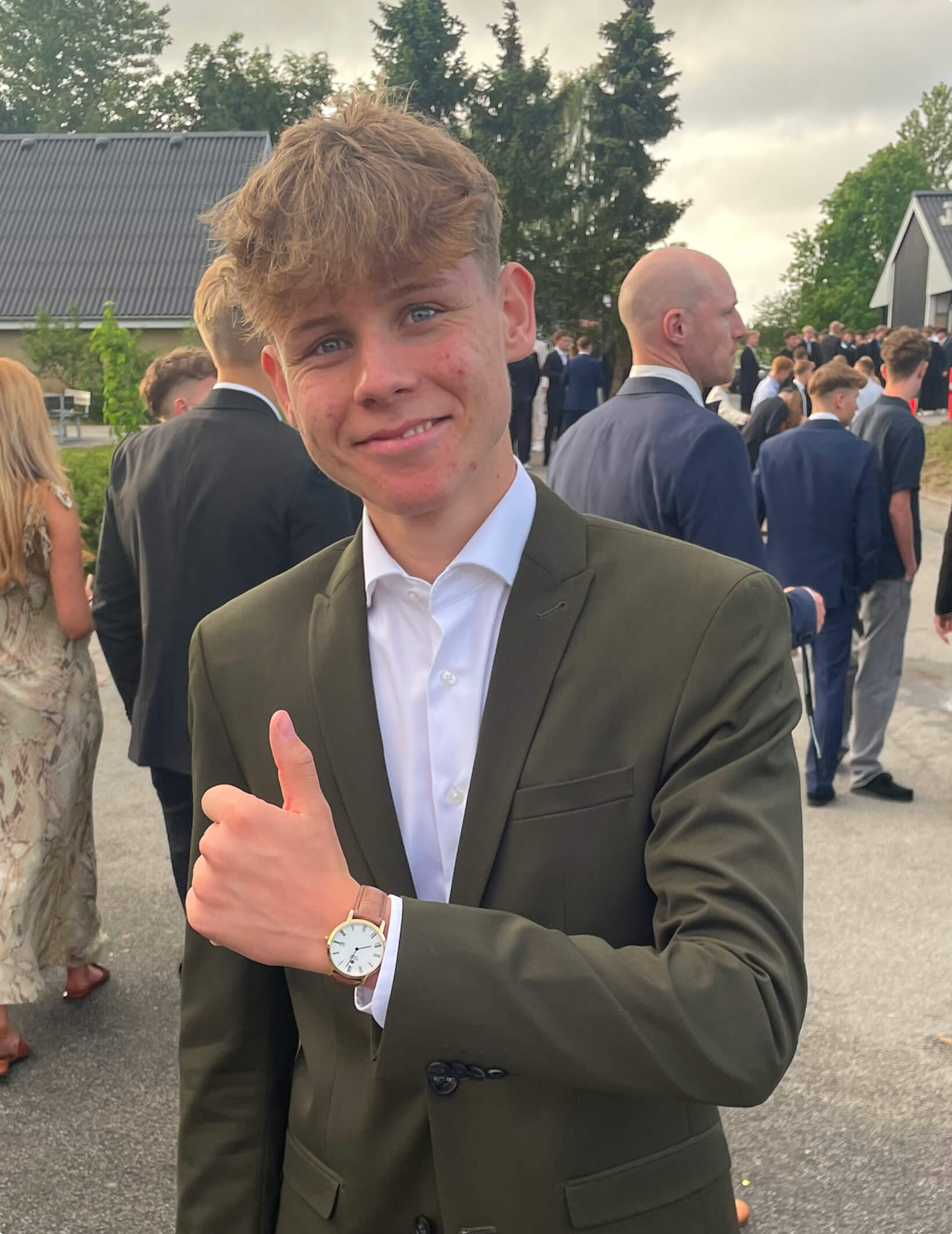 A young man in a green blazer smiles and gives a thumbs up, with people in formal attire in the background.
