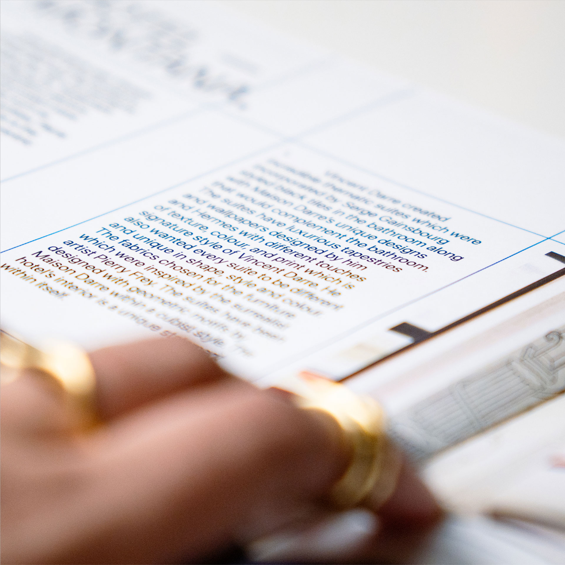 A hand with gold rings gracefully turns the page of a magazine, showcasing a close-up of blue printed text and blurred images, emphasizing the tactile experience of reading.