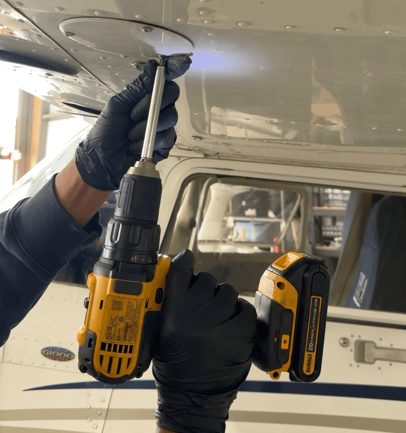 A technician wearing black gloves uses a yellow cordless power drill to remove screws from an access panel on the underside of an aircraft fuselage