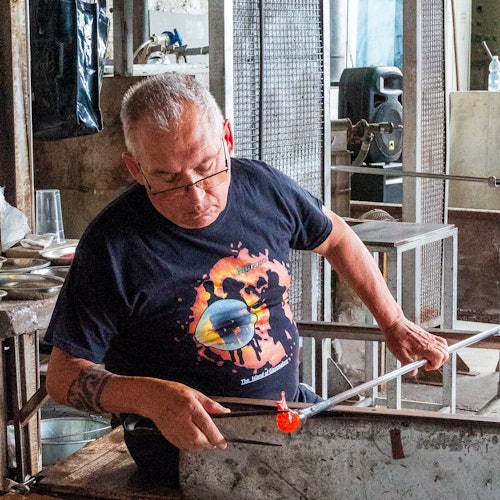 一位戴着眼镜、穿着黑色T恤的工匠正在工坊里用一根杆子塑形一块发光的玻璃。