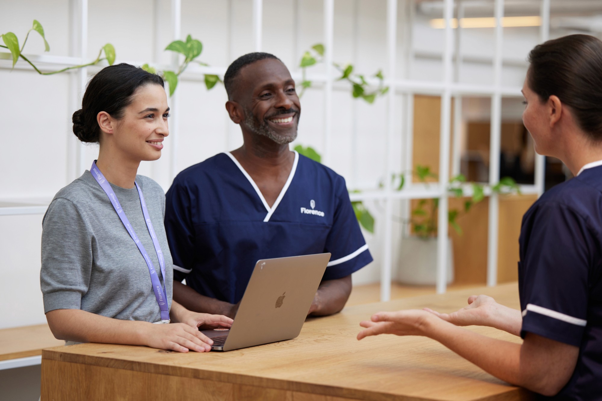 Three healthcare workers having a conversation at a reception desk, including a man in a Florence-branded navy nursing uniform holding a laptop