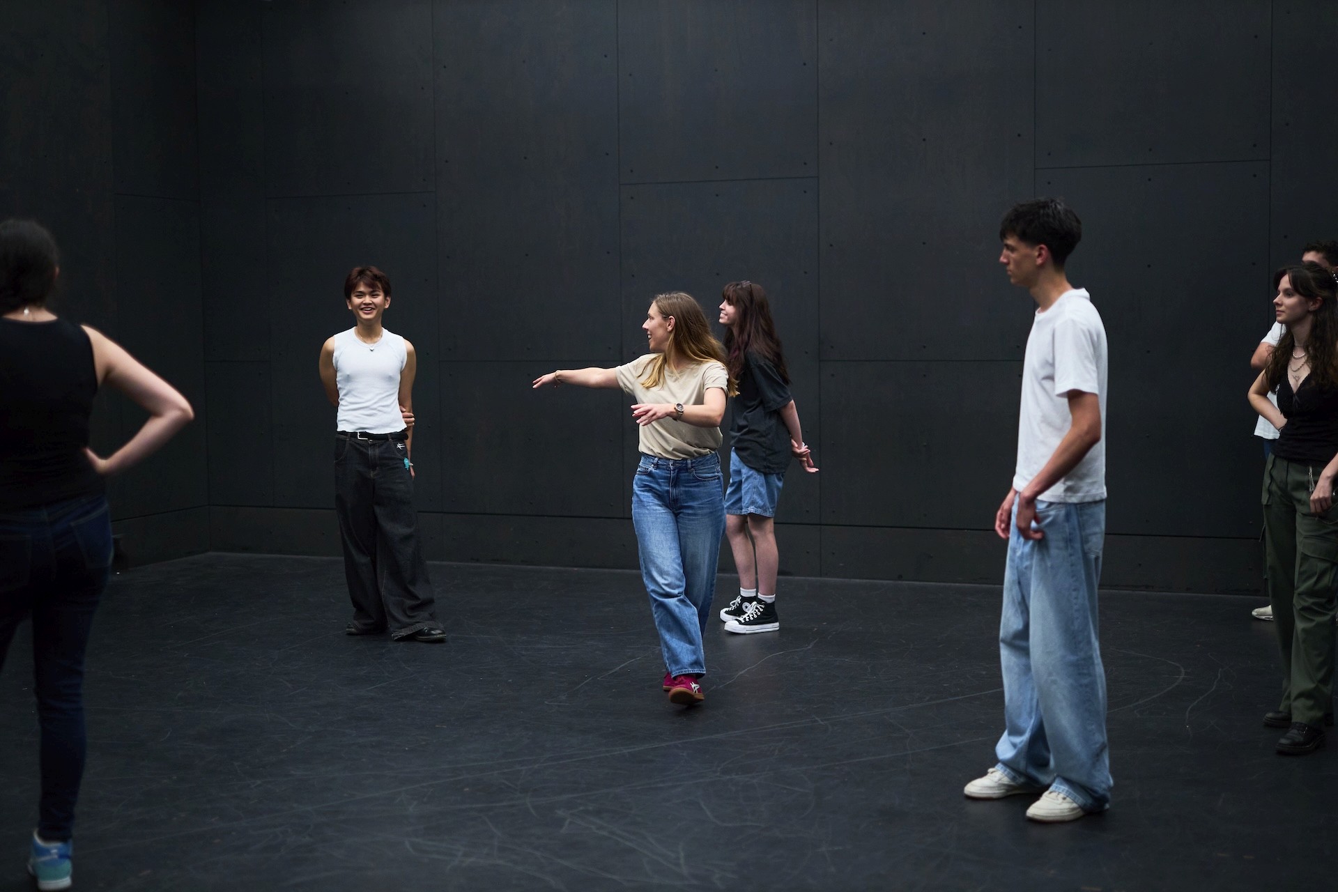 A child stands with one arm raised while other children lie on the floor around her during a drama game.