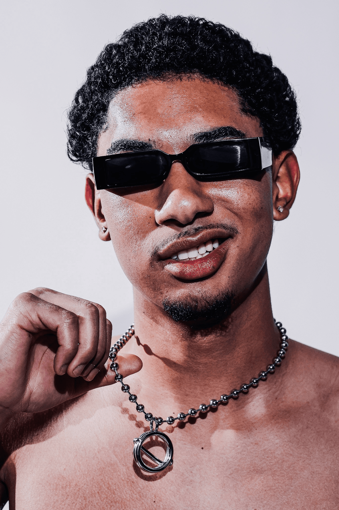 A man with curly hair, wearing black sunglasses, smiles confidently while holding a silver ball chain necklace with a circular pendant. He is shirtless, and the focus is on the close-up of his face and upper body, set against a plain light background.