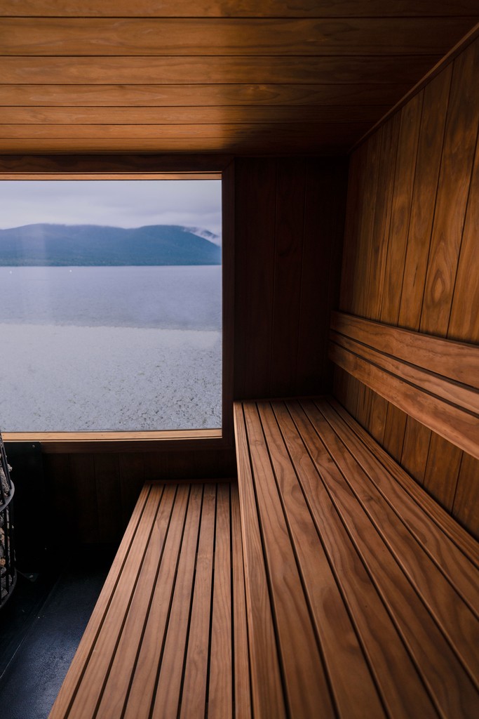 A wooden door stands open by a serene lake, reflecting the overcast sky and distant mountains. The scene is calm and introspective, with muted tones.