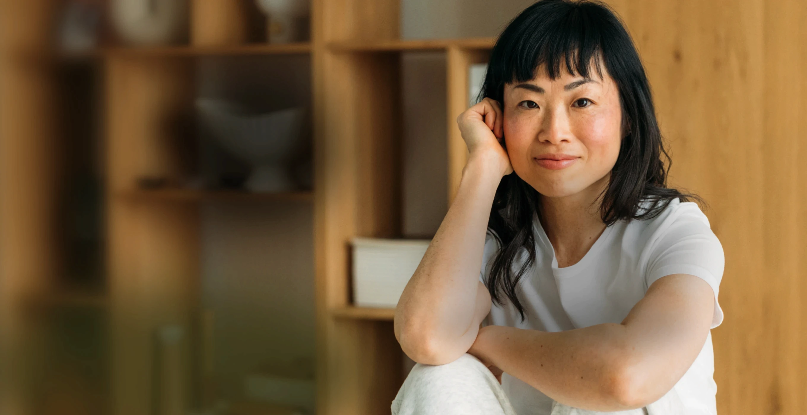 A woman named Jenny O with black hair smiles while sitting on a wooden surface, with shelves in the background.