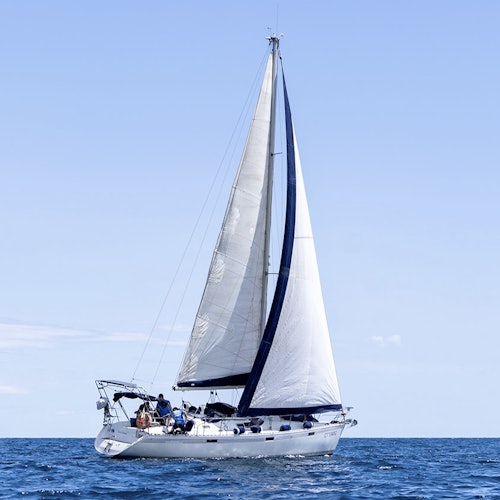 A sailboat with white sails navigates on a calm ocean under a clear blue sky; people are visible on the boat's deck.