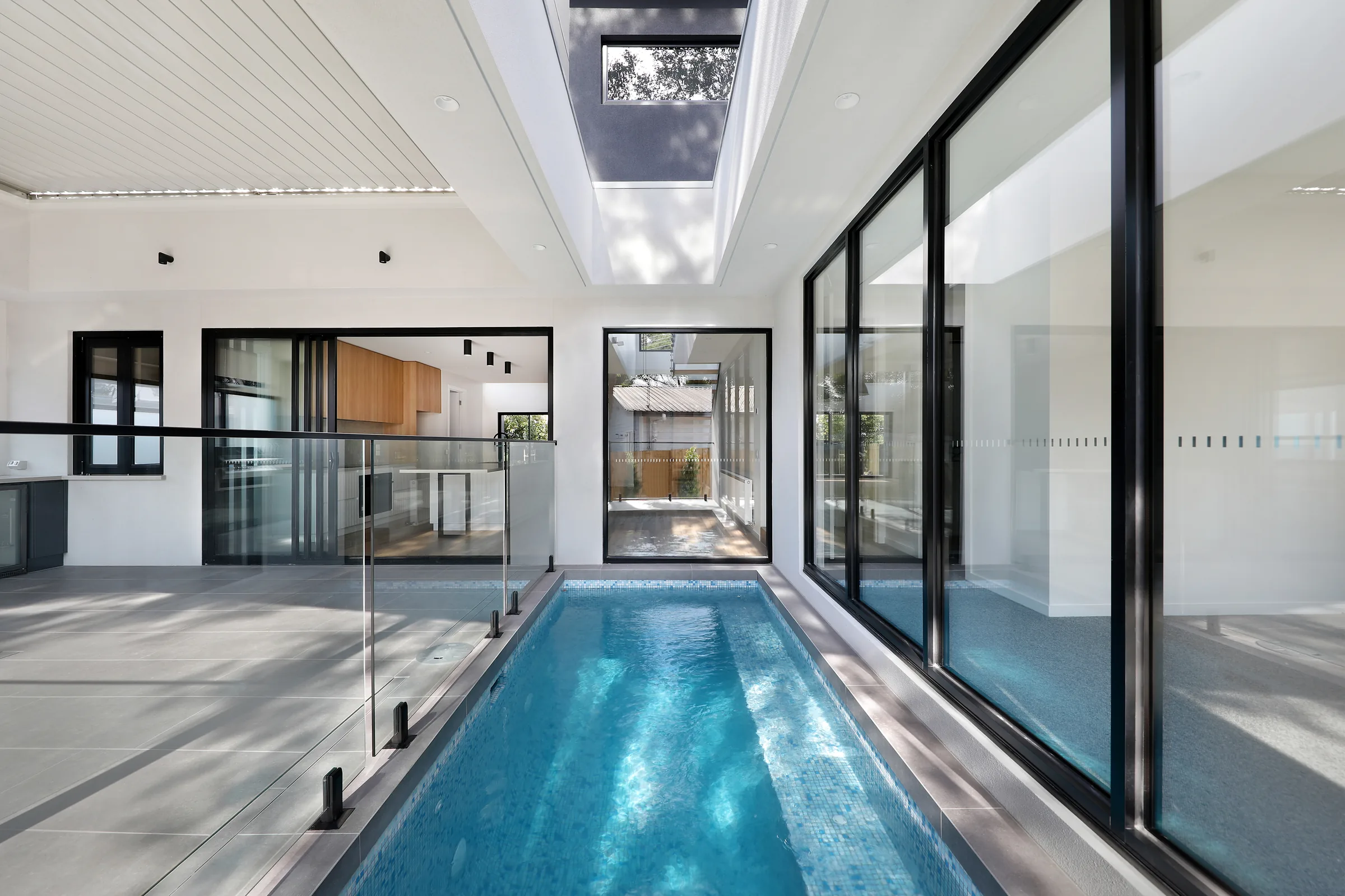 Internal lap pool in blue mosaic tiles beneath a large skylight, framed by black-framed glazing and a glass balustrade, with grey tiled surrounds and a glimpse of the timber kitchen
