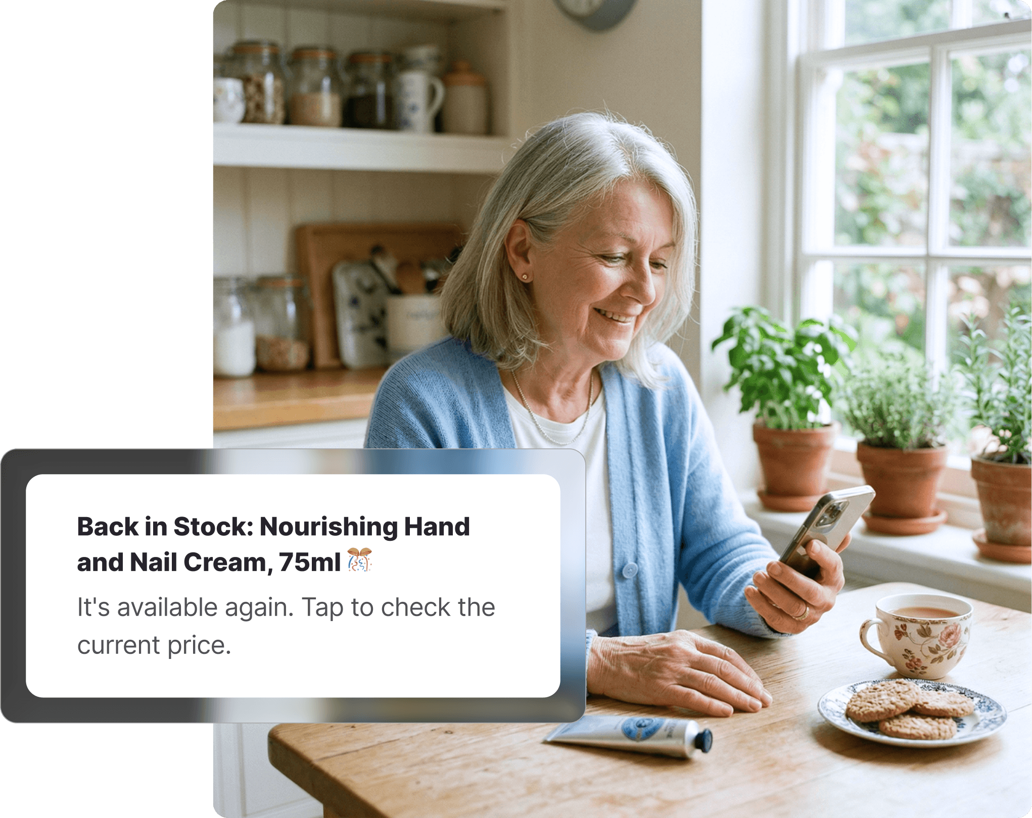 Older woman sitting at a kitchen table smiling at her phone, with a Priceline-style hand cream on the table beside a cup of tea and biscuits, receiving a back-in-stock notification on Whisprice.
