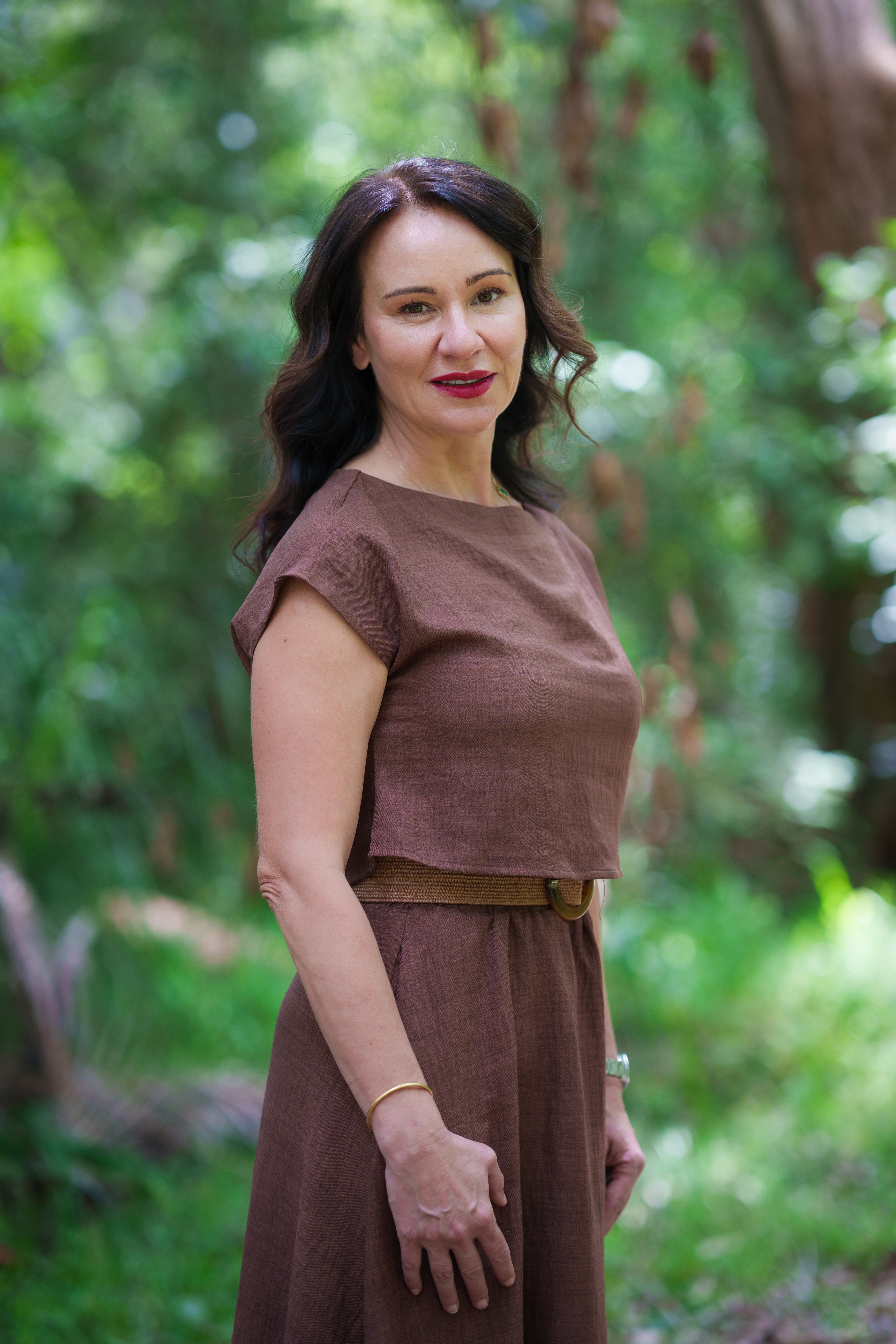 Dr. Bonnie Hawthorne with dark hair stands in a lush green setting, wearing a brown dress and smiling softly.