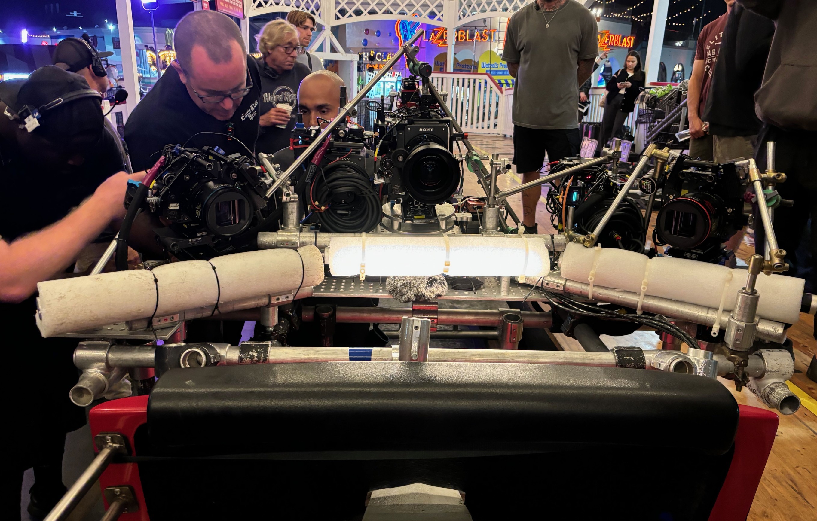 Three Sony cameras and an LED light panel mounted on a speedrail and cheeseplate rig on a roller coaster car at Belmont Park during a night shoot, with foam vibration padding visible and film crew surrounding the setup