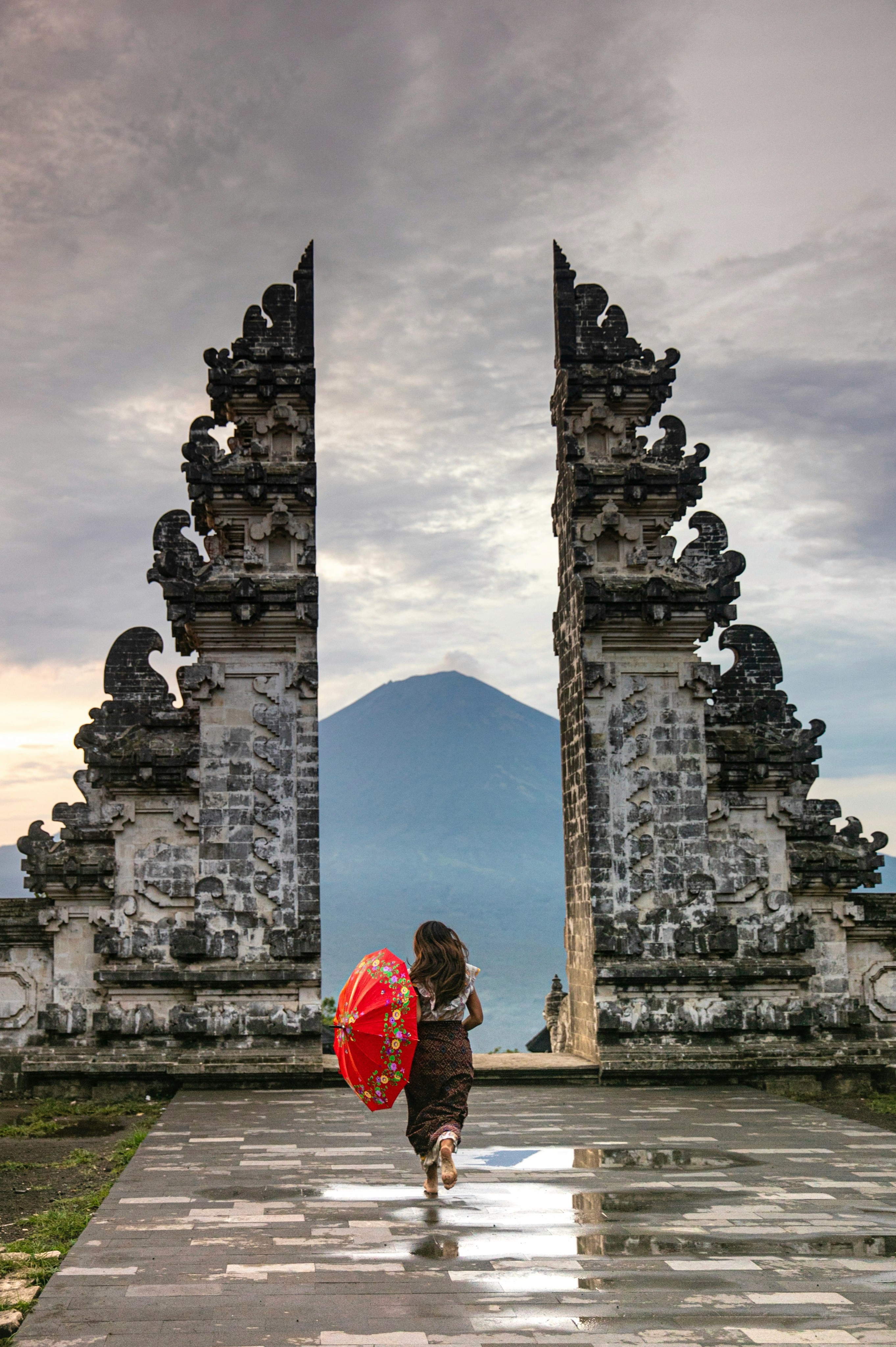 A person with a red umbrella walks through a temple gate toward a mountain.