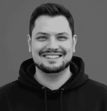 Woman in sunglasses poses against vibrant red background, wearing a beige blazer and black top. Sunlight casts dynamic shadows, creating a stylish, confident mood.