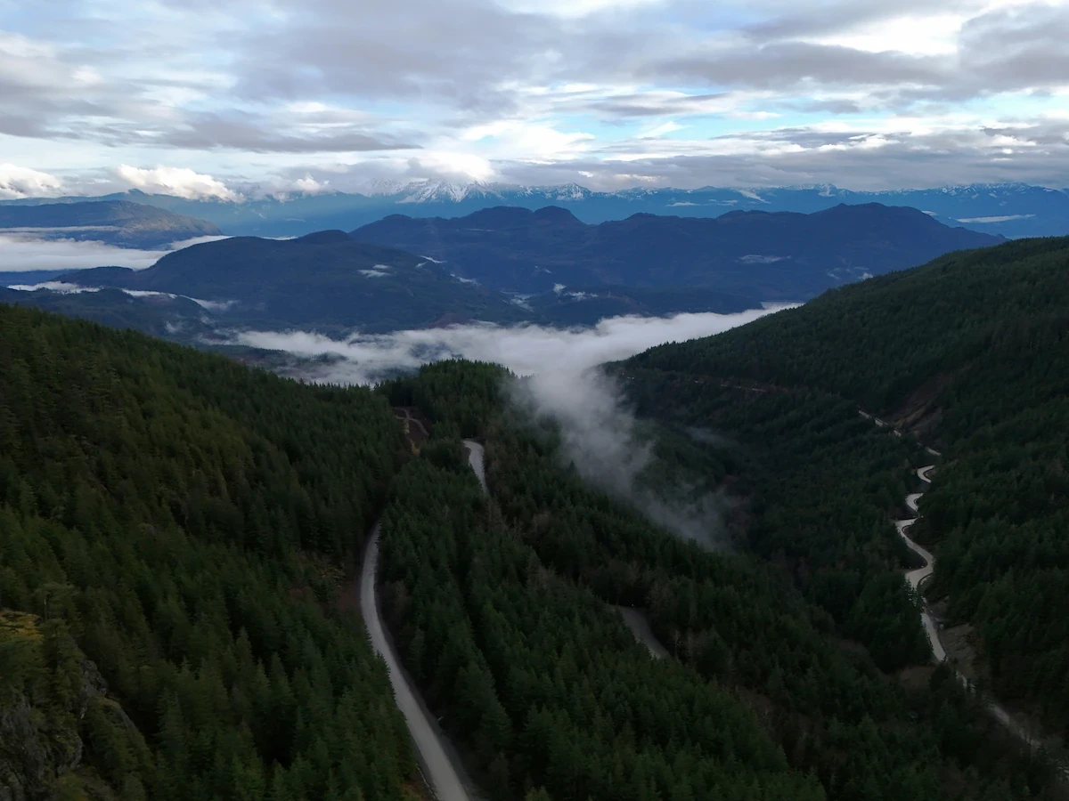 Aerial view of the forested mountains surrounding Agassiz, BC with a winding road and low clouds drifting through the valley.