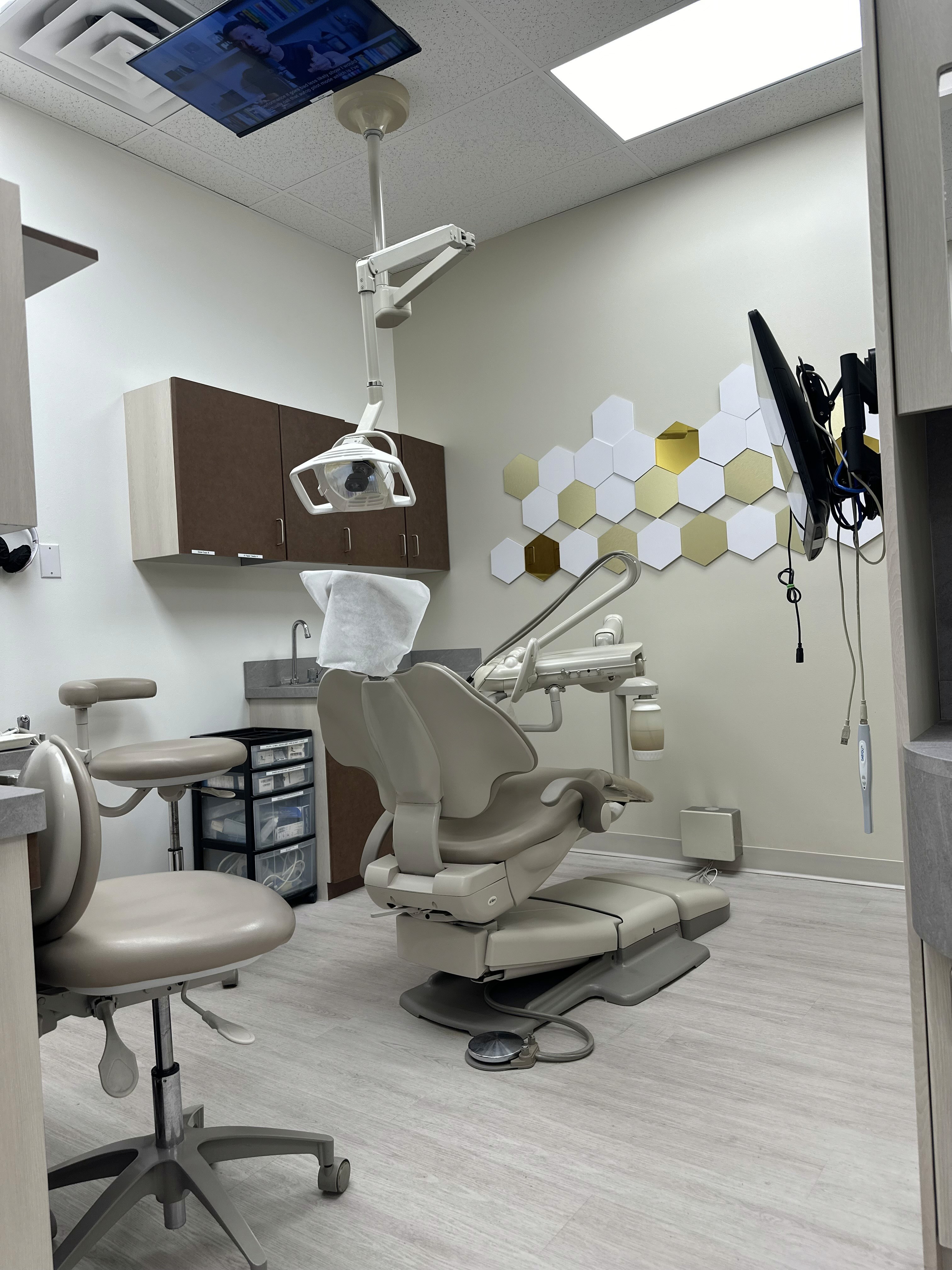 A modern dental treatment room featuring a dental chair, overhead light, dental equipment, and cabinets, with hexagonal wall decor in the background.