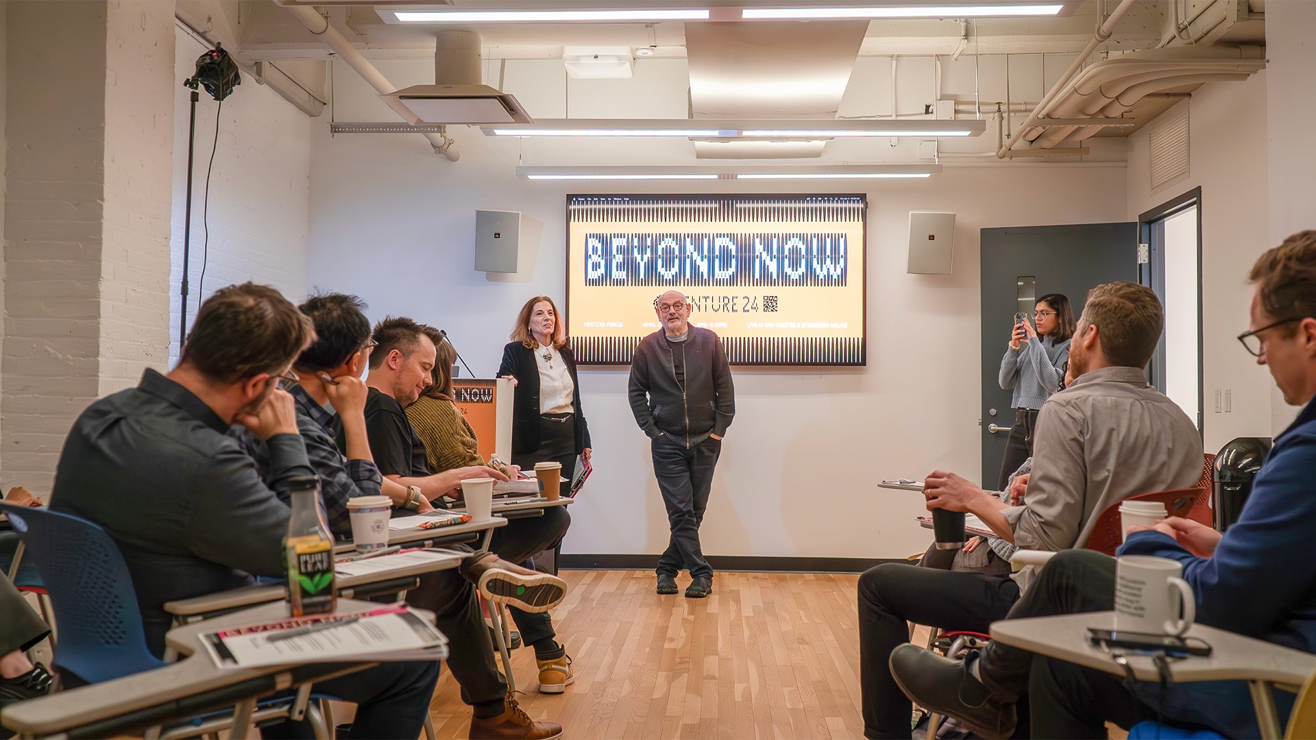 Two people speaking to a crowded room, other people seated at desks.