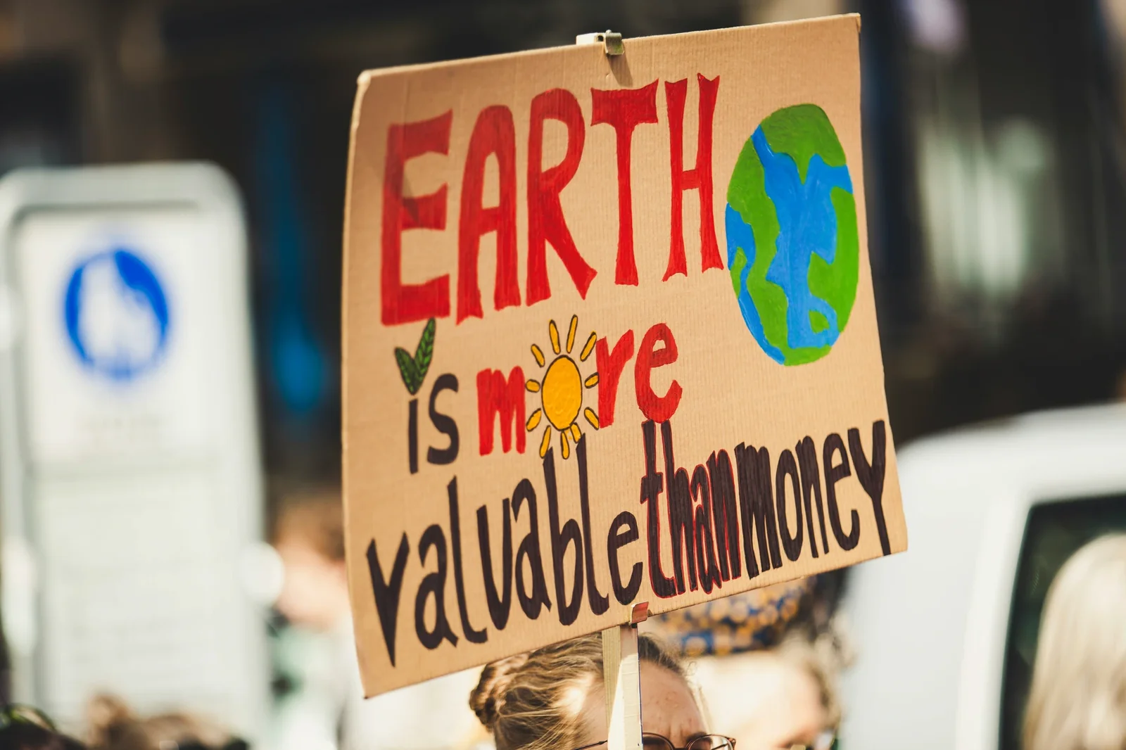 Protest sign reading "EARTH is more valuable than money" with painted globe and sun, held at sustainability demonstration.