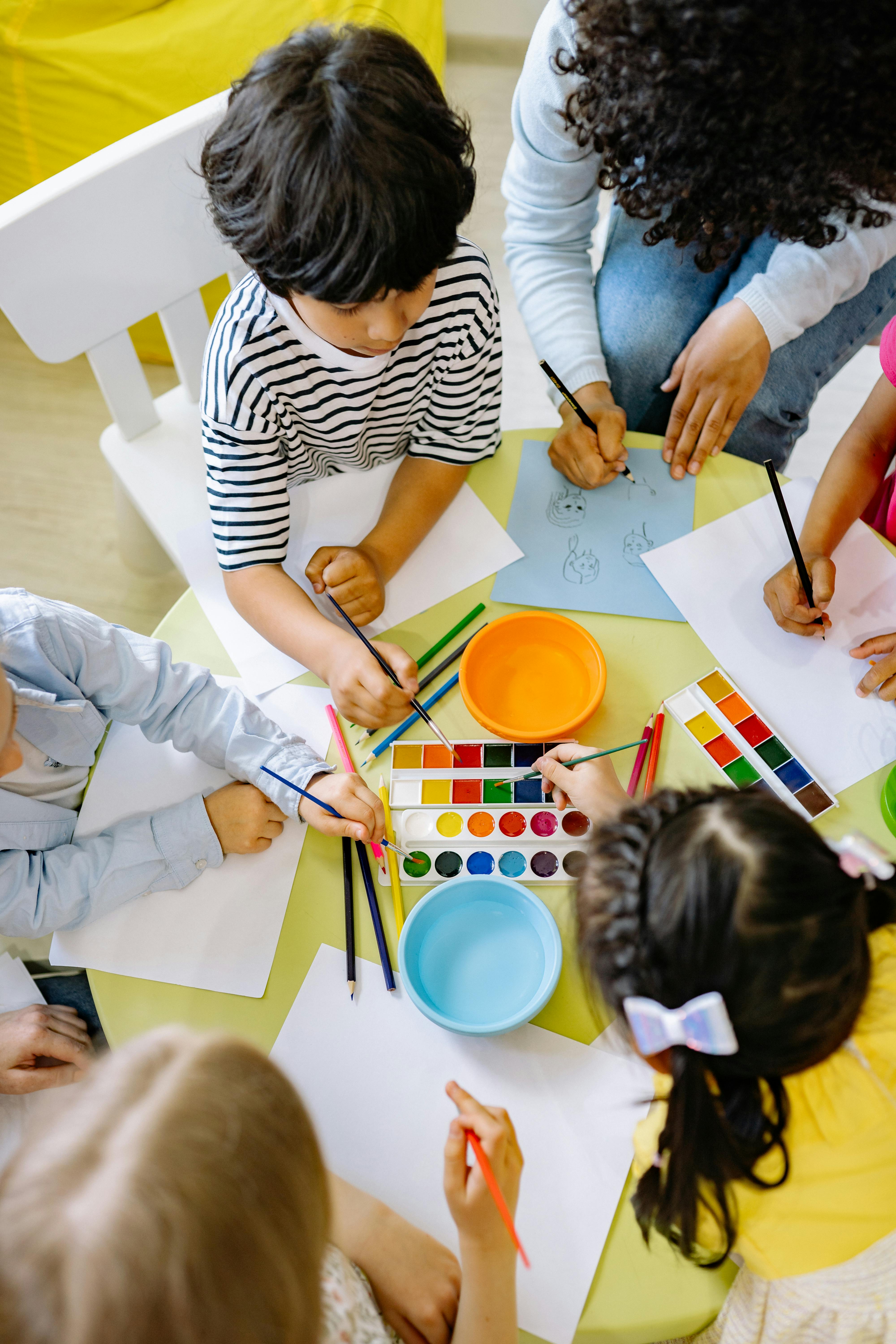 Vista aérea de un grupo de niños y niñas de diversas edades sentados alrededor de una mesa amarilla participando en una clase de arte o taller de arteterapia inclusivo en San Fernando. Usan acuarelas y lápices de colores bajo la guía de una docente en Espacio Yutori.