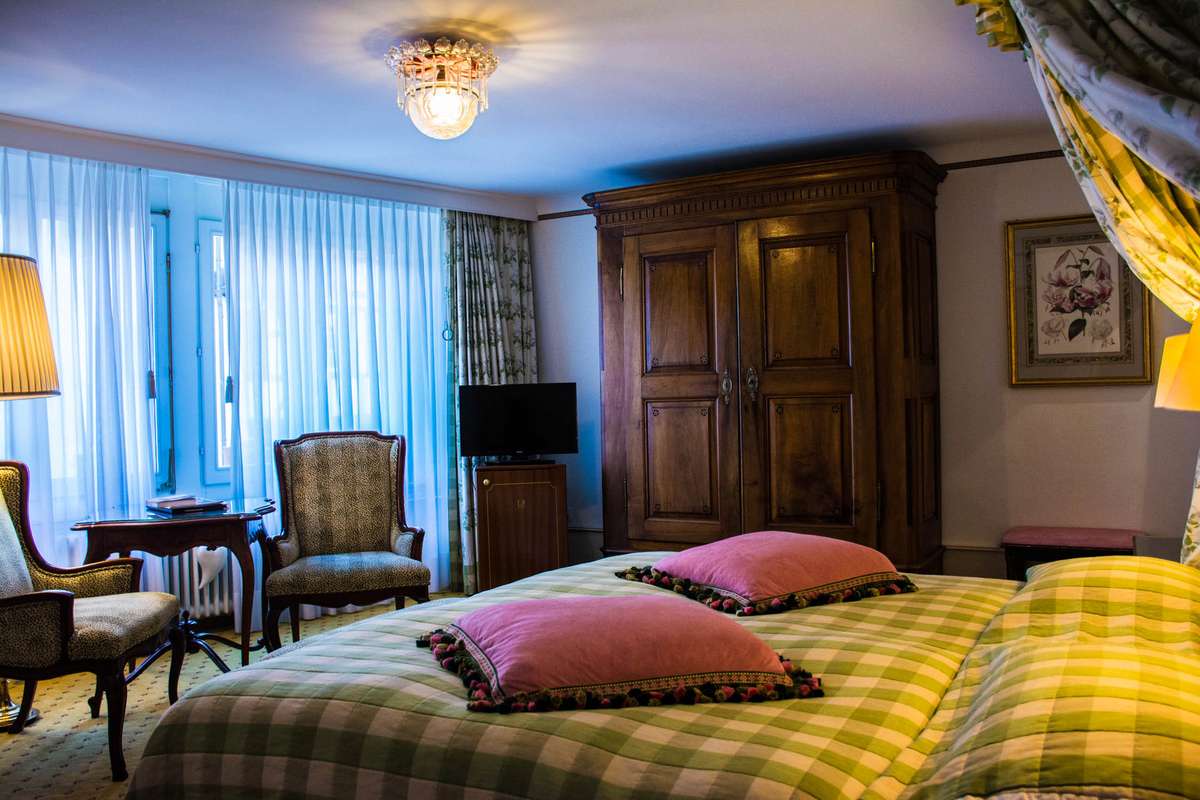 A hotel room with a checkered bedspread, two pink pillows, a wooden wardrobe, and a large window with sheer curtains.