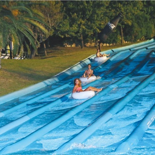 Three people in inflatable tubes sliding down a multi-lane water slide, surrounded by grass and trees.