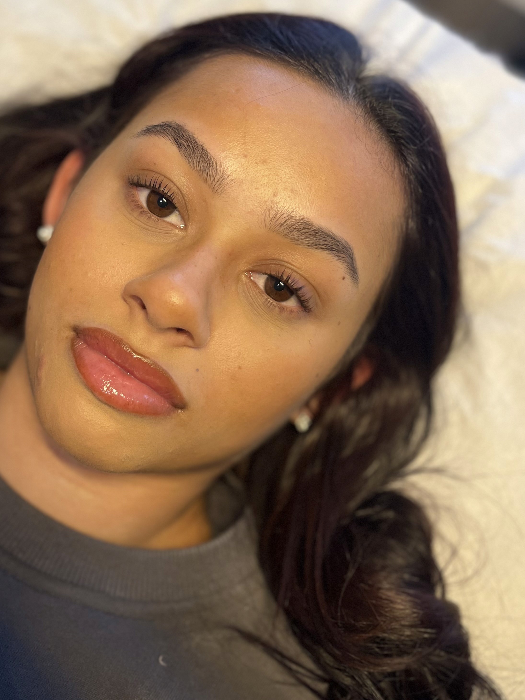 Woman with long dark hair, lying down on a white surface. Her expression is calm, and she wears a gray top with shiny, natural makeup.