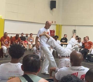 Zwei Capoeira-Spieler in einer Roda, einer führt einen hohen Kopftritt aus, während der andere ausweicht, umringt von sitzenden Zuschauern und Musikern.