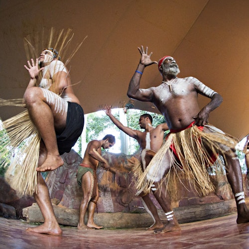 Men in traditional attire performing a cultural dance, with grass skirts and body paint, on a wooden stage.