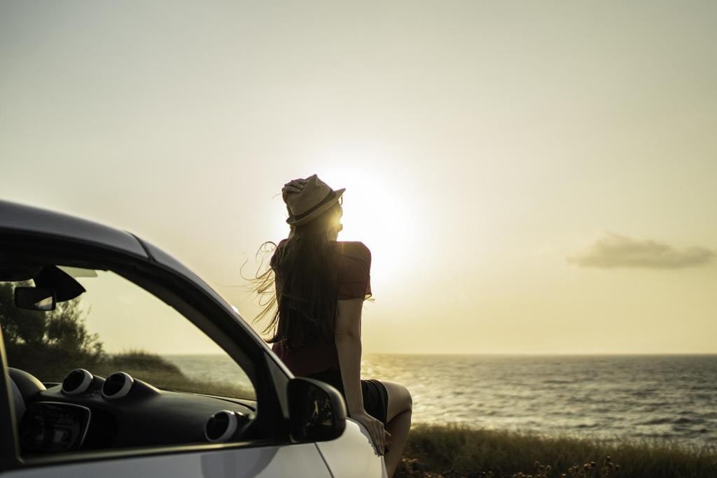 Silhouet van een persoon bij het strand bij zonsondergang, leunend tegen een auto, met zachte golven op de achtergrond.