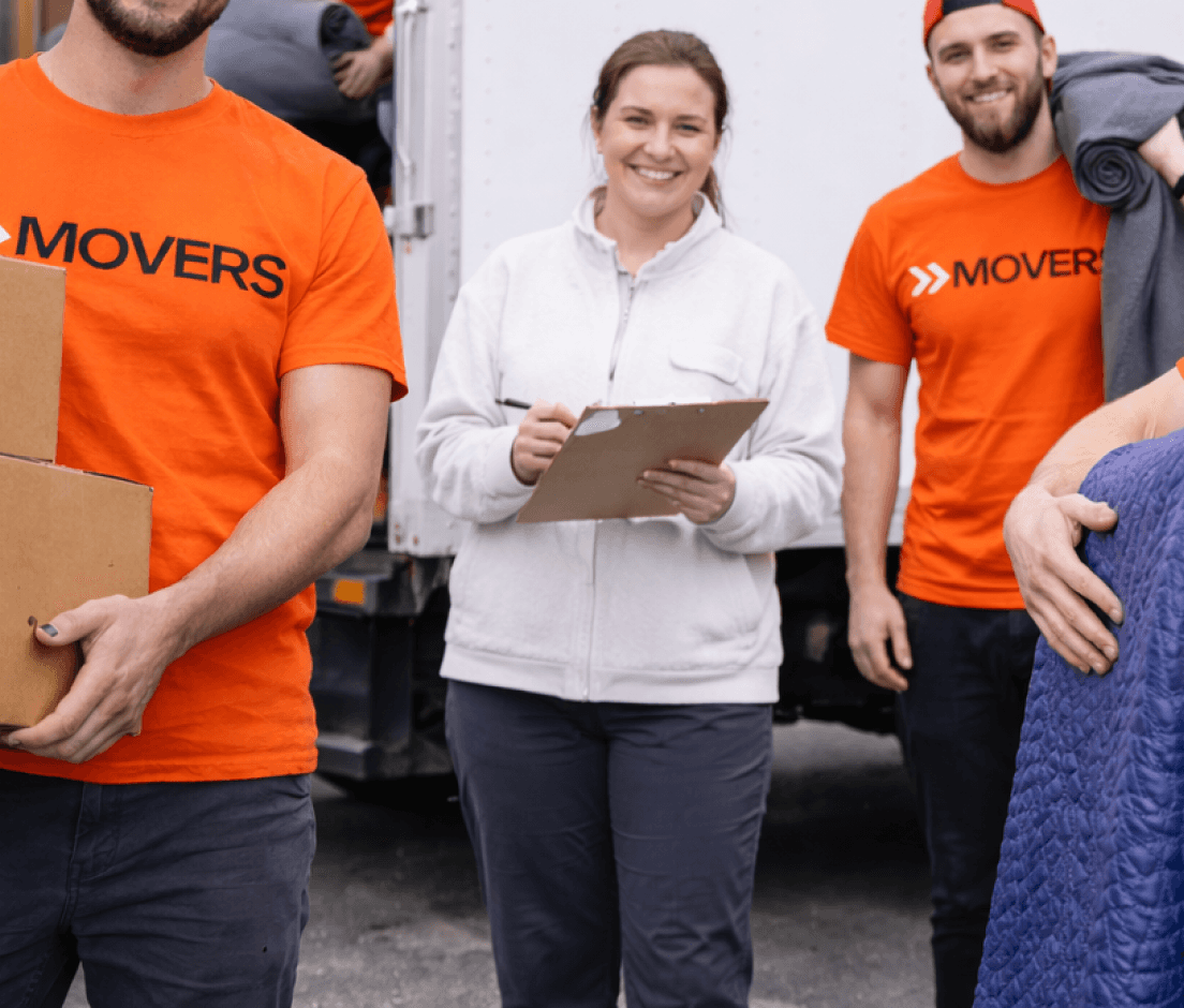 Smiling movers in orange shirts and a supervisor holding a clipboard stand in front of a white moving truck.