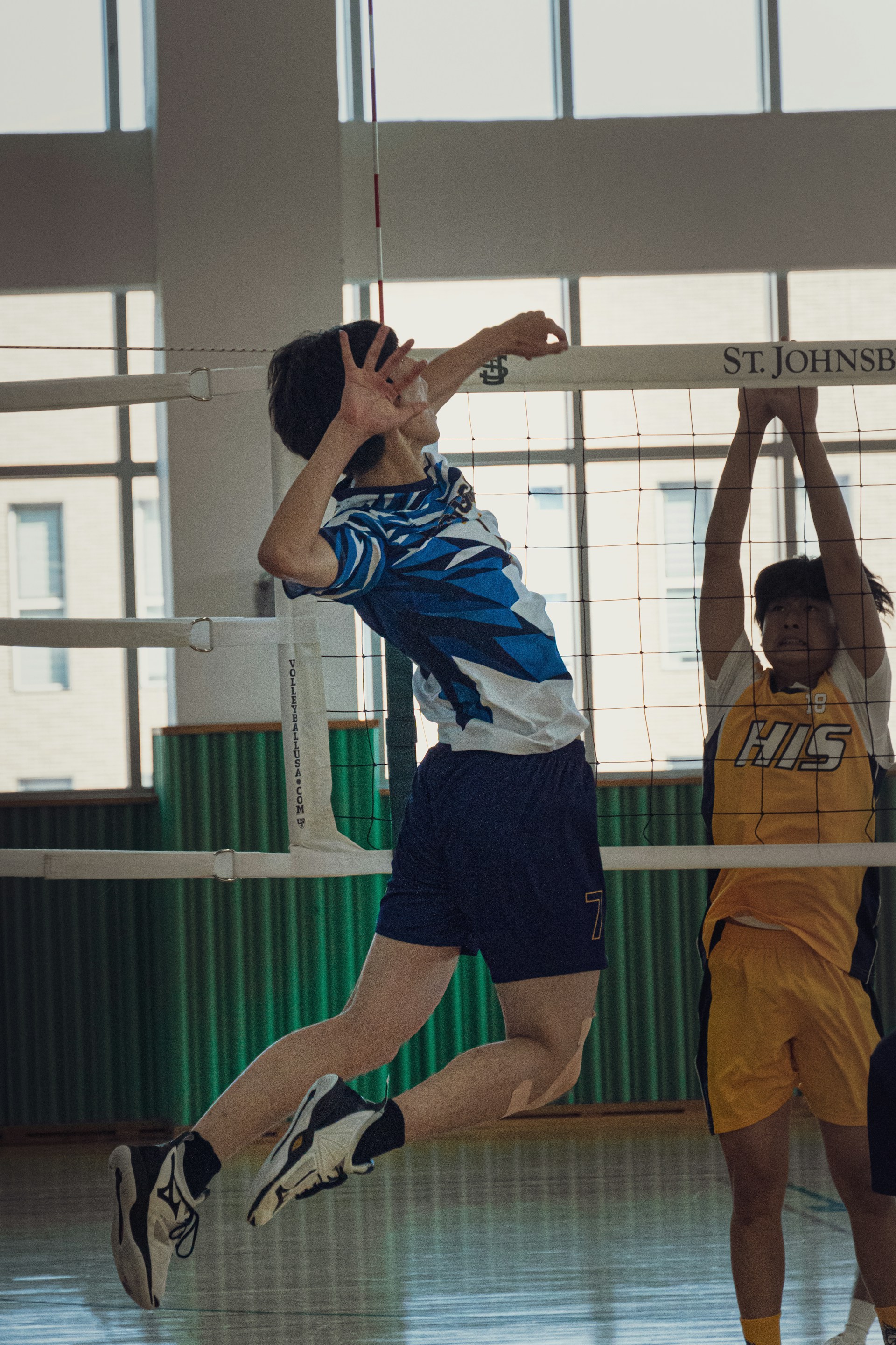 A volleyball player in a blue and white patterned jersey spiking the ball past a blocker in a yellow "HIS" jersey.