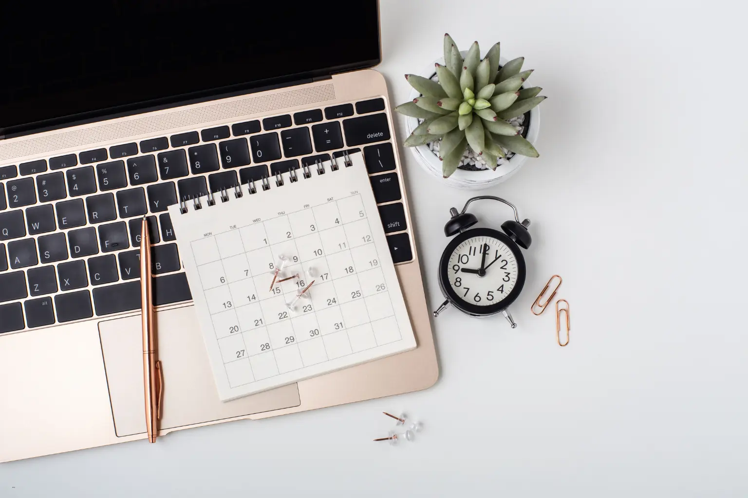 Automated calendar scheduling setup with a laptop, desk calendar, alarm clock, pen, and succulent plant on a workspace