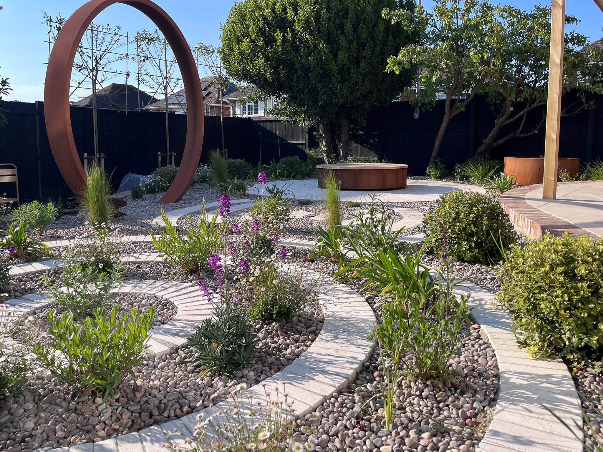 A landscaped garden featuring curved pathways, circular structures, and various plants under a clear blue sky.
