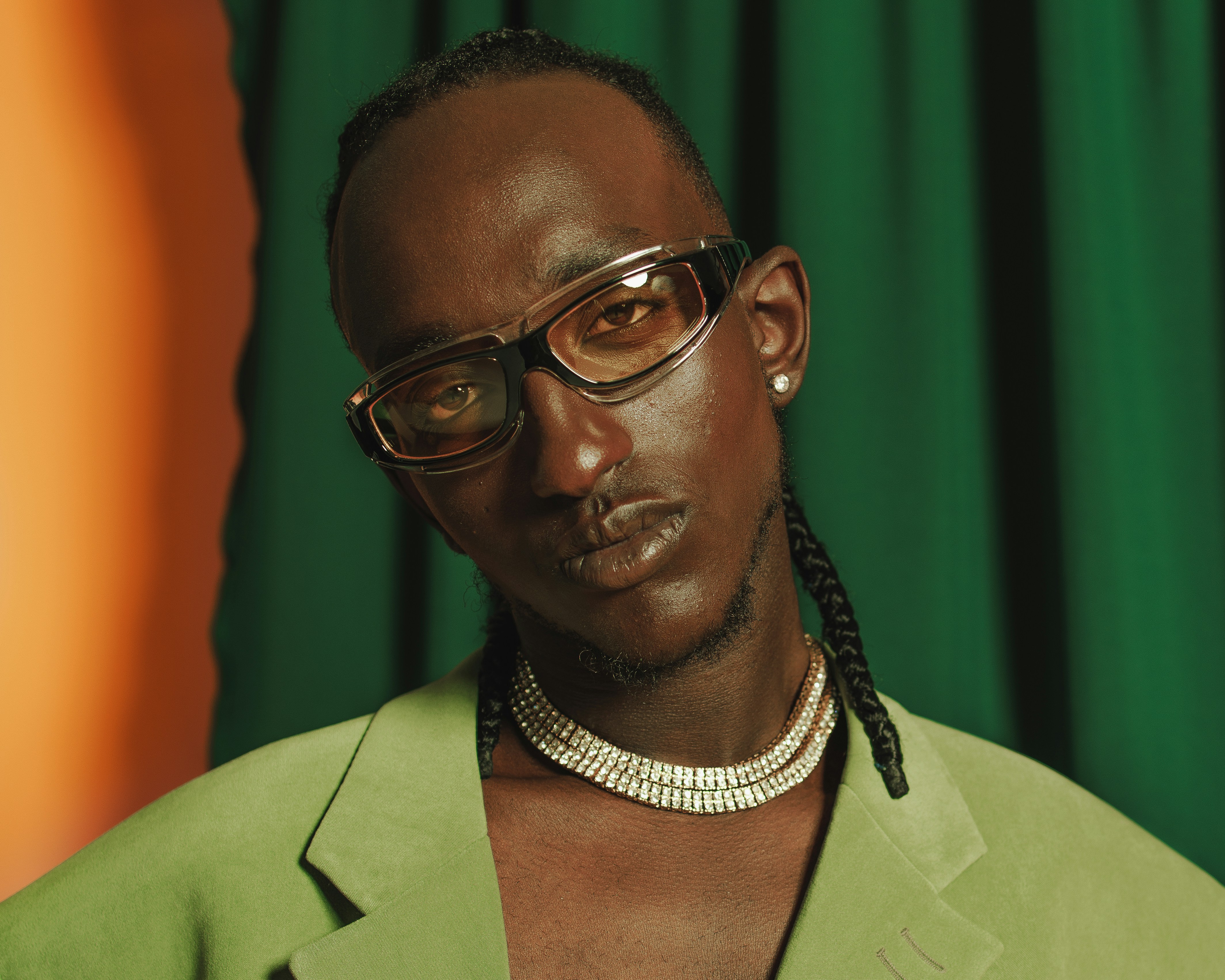 A man poses wearing vibrant green clothing in front of a green curtain and orange wall