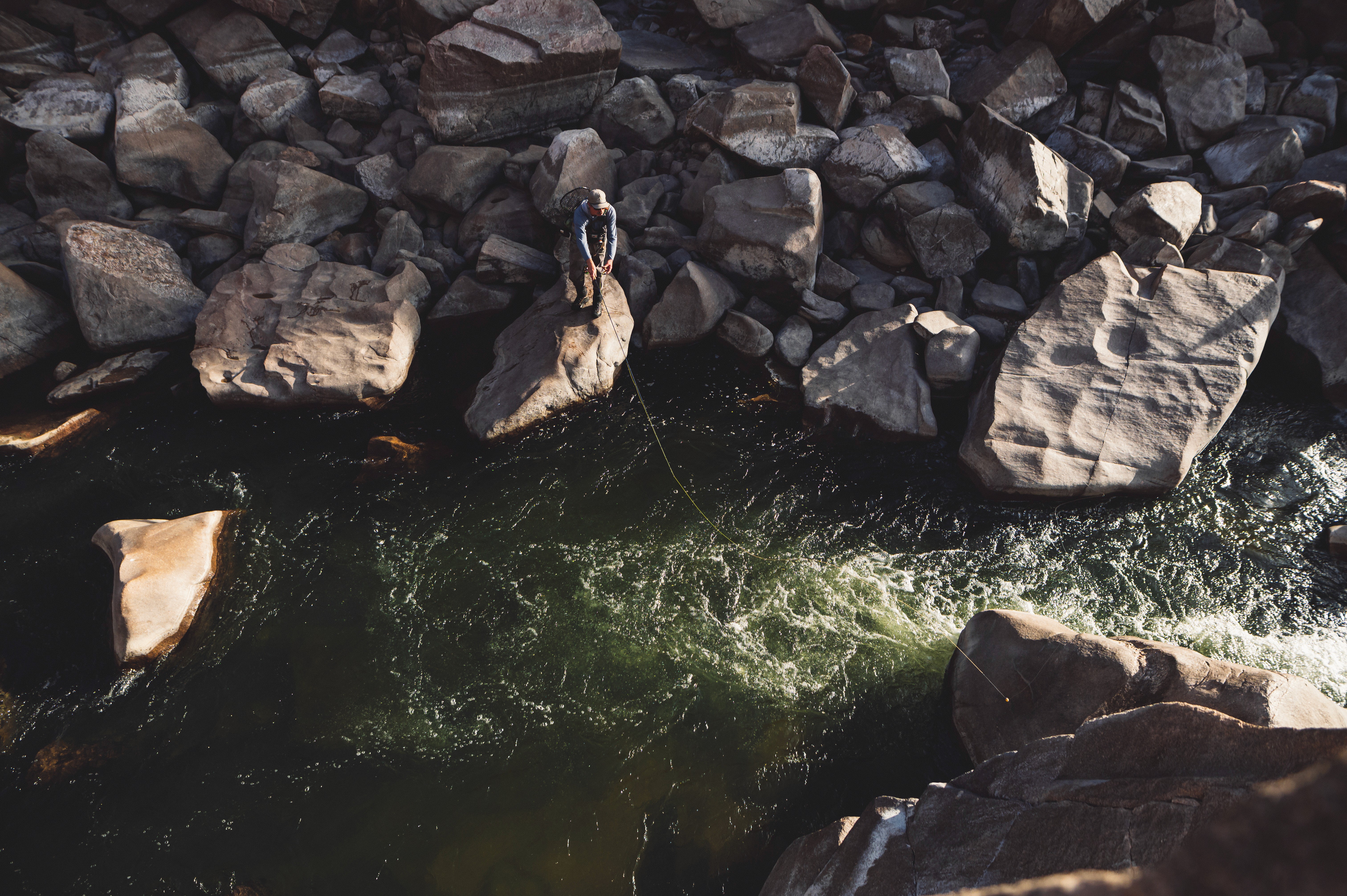 man fly fishing off rocks into a flowing river. large bolders drone shot