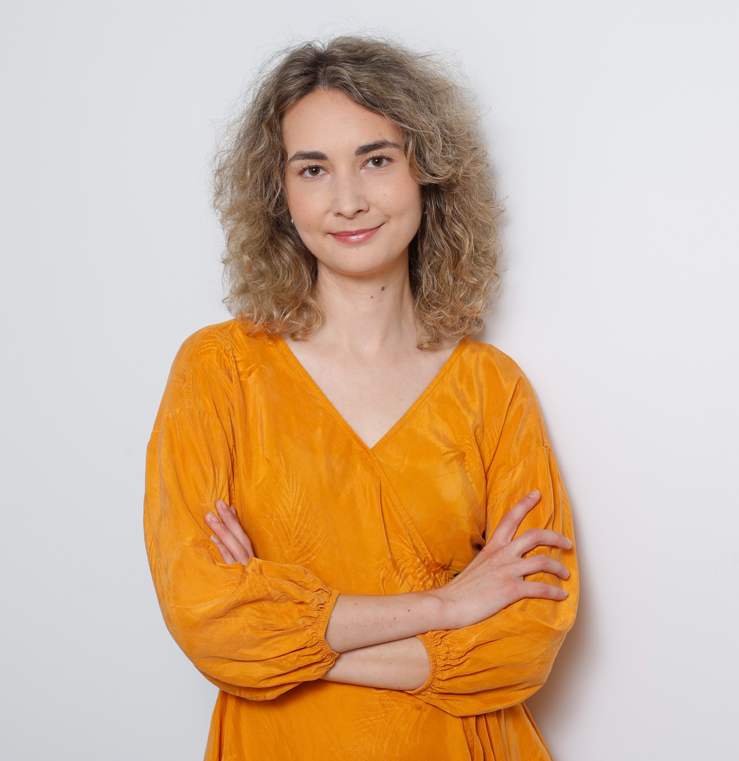 Smiling female therapist with short gray hair, wearing a brown top and a purple shawl, surrounded by a cozy background.