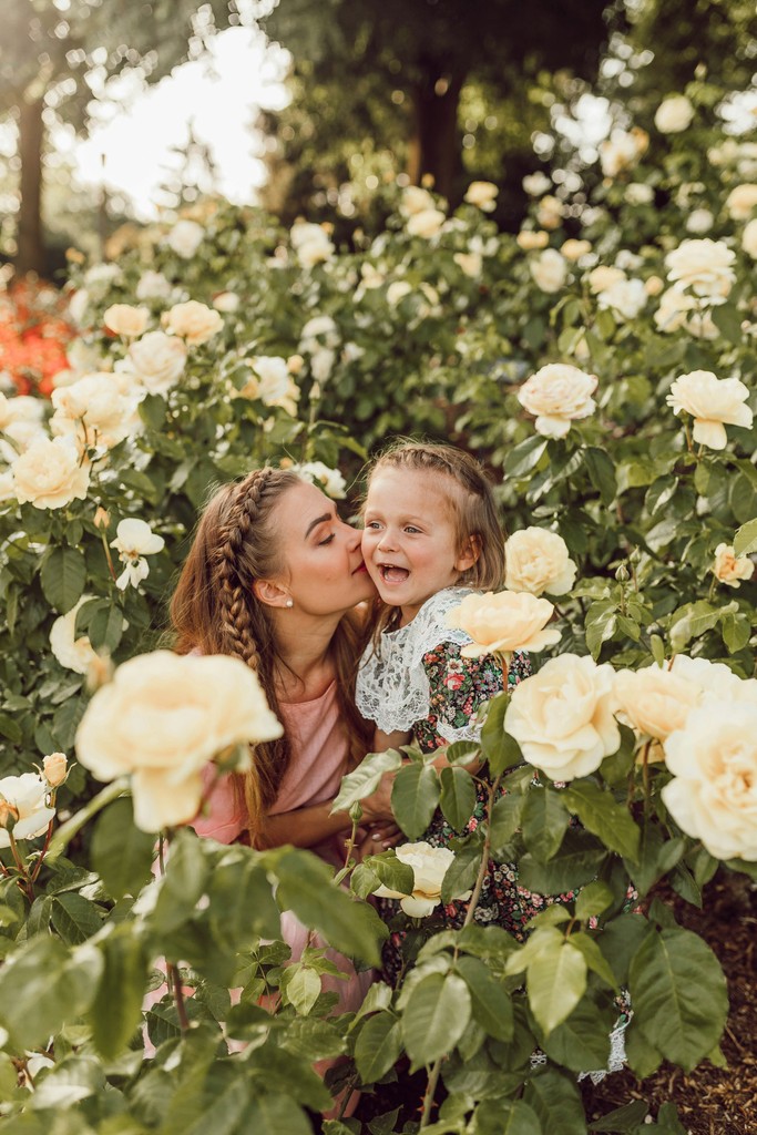 A mother and her young daughter share a tender moment in a lush garden filled with blooming yellow roses. The mother gently kisses her daughter's cheek as they both smile, surrounded by vibrant greenery and flowers. The scene captures the beauty of their bond and the joy of spending time together in nature.