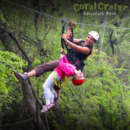 两个人在森林中体验滑索项目，一名成年人支持着一个倒挂的孩子。文字："珊瑚坑冒险乐园。"