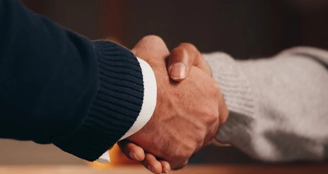 Duas pessoas apertando as mãos sobre uma mesa de madeira, uma de blazer escuro e outra de suéter claro