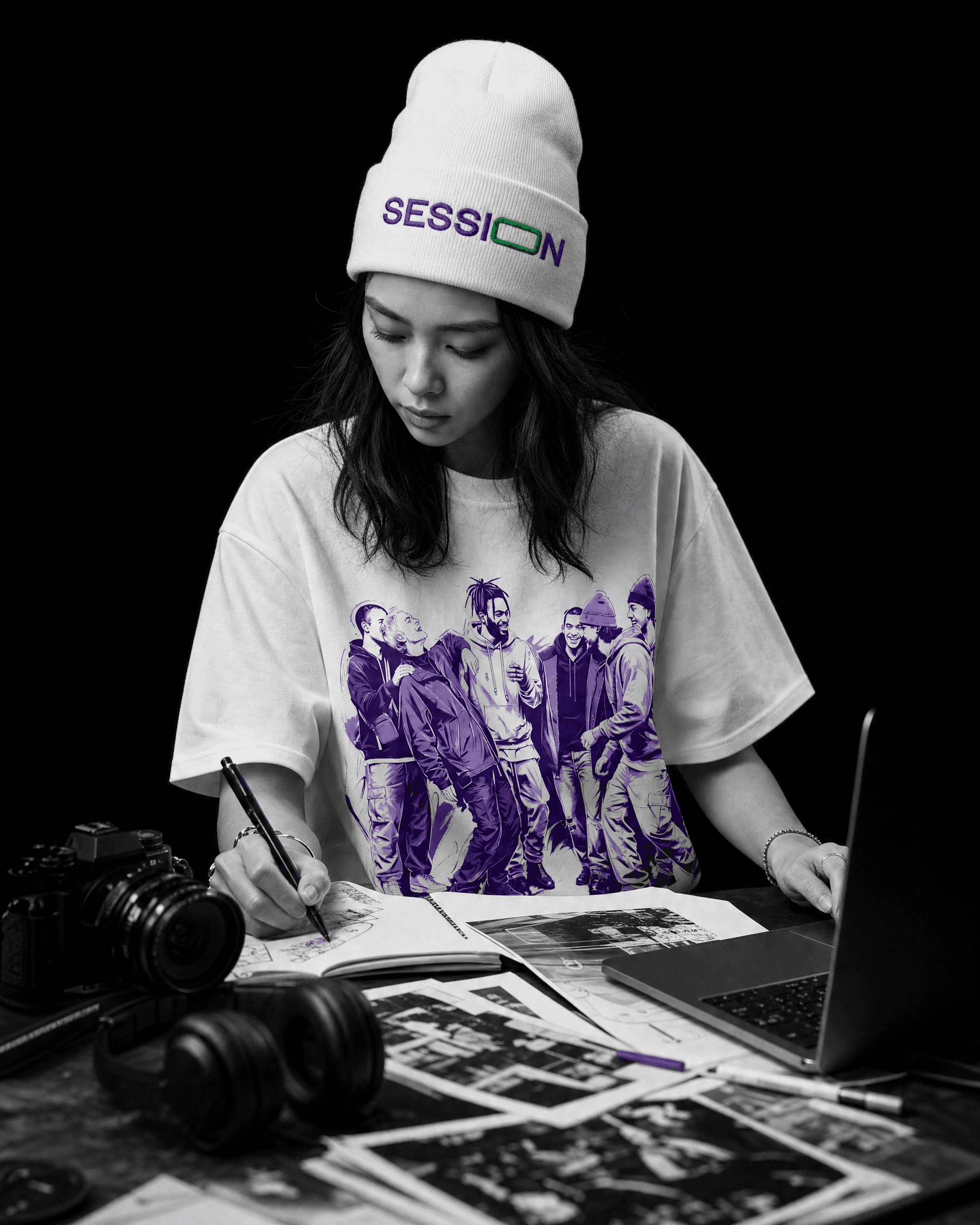 Woman in a white Session Zone beanie and community-graphic T-shirt sketching beside a laptop and camera.