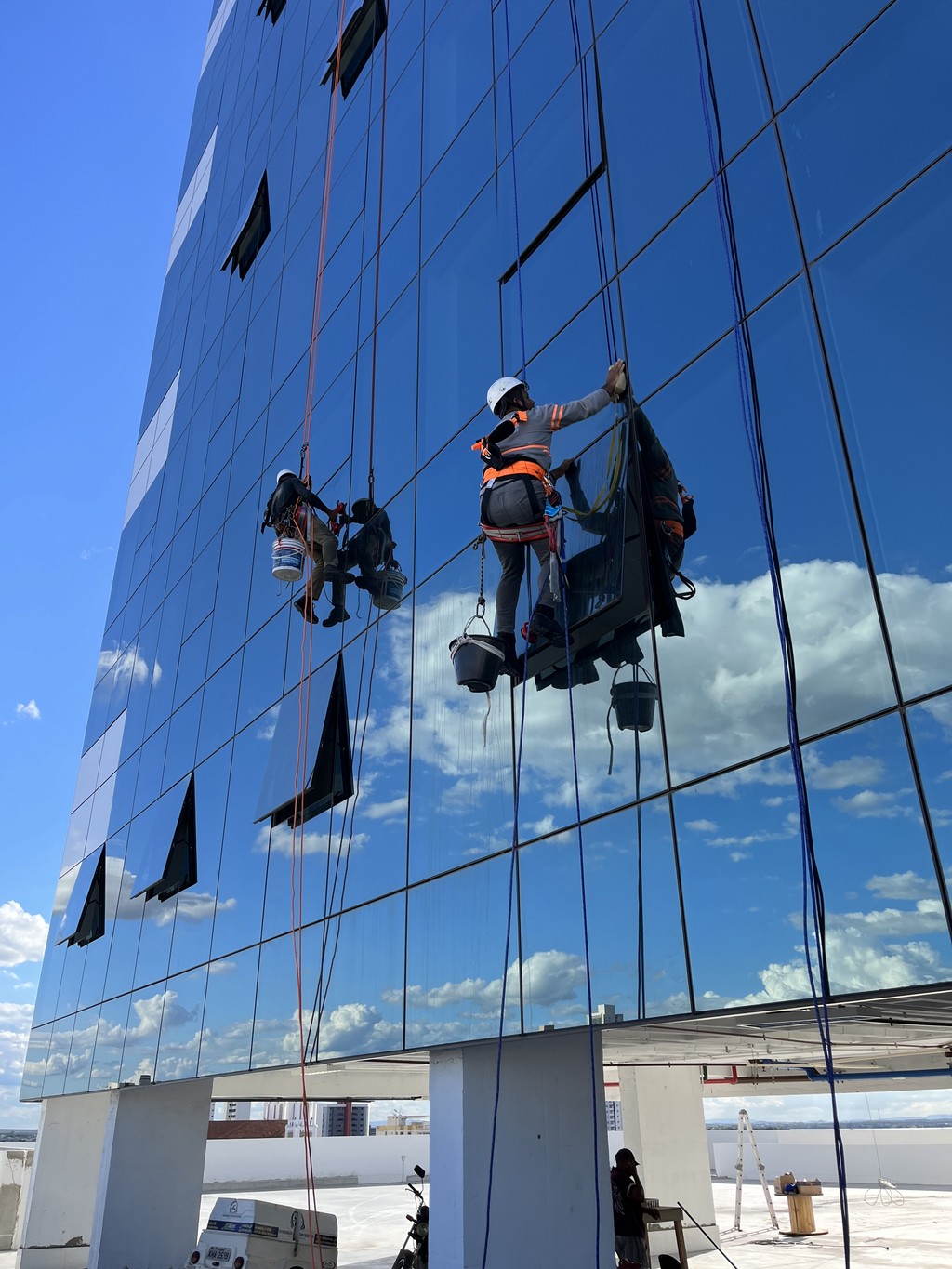 Equipe de manutenção predial realizando limpeza de fachada de vidro do Trade Center Dom Campelo, dois profissionais equipados com EPIs de segurança laranja suspensos por cabos e cadeiras balanceiras contra céu azul com nuvens, demonstrando cuidado com manutenção e segurança do edifício.